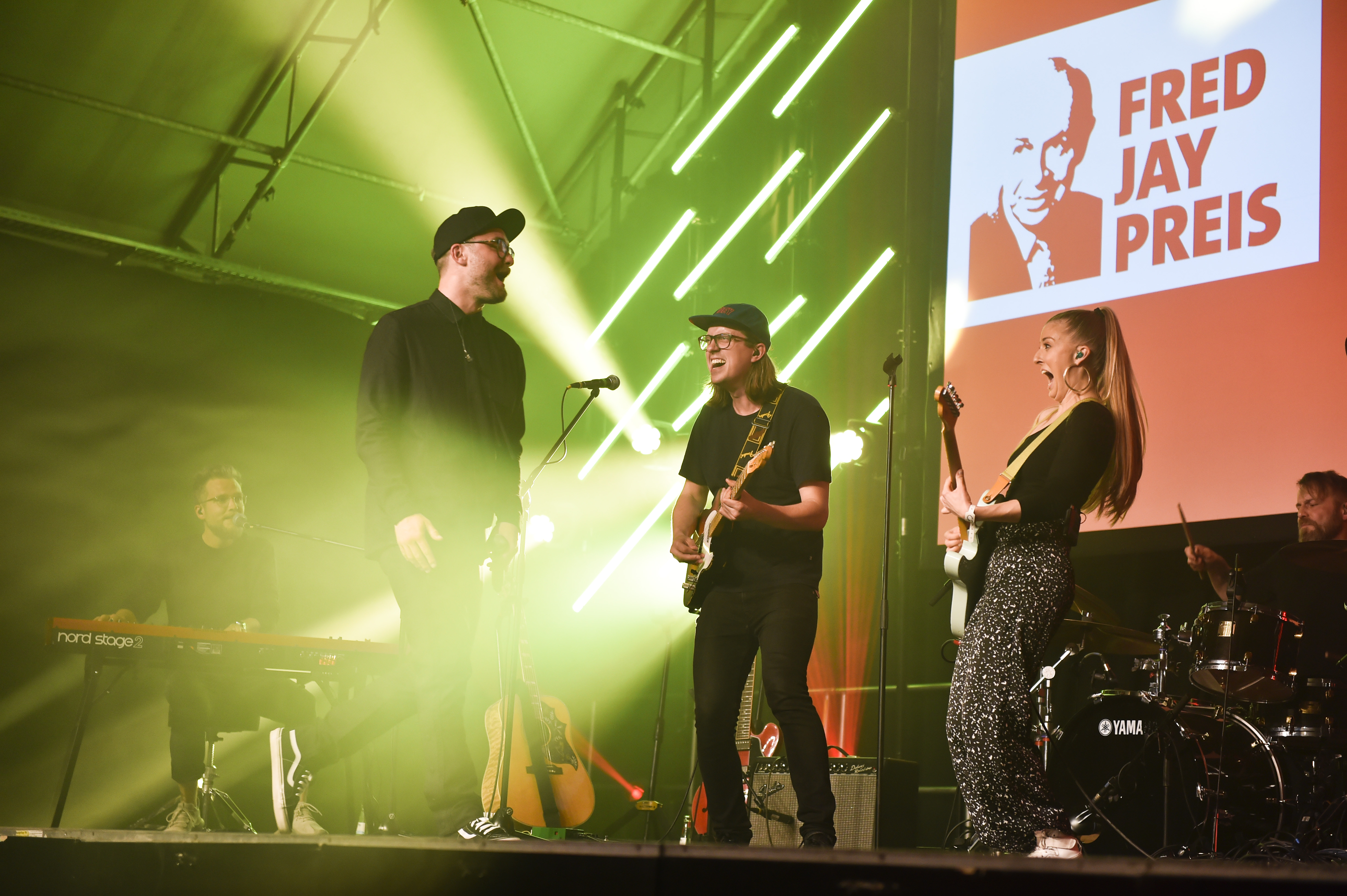 Band performing energetically on stage with guitars and drums under green lights, 