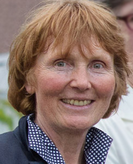 A person with short, light brown hair smiles outdoors, wearing a dark jacket and a polka dot shirt.