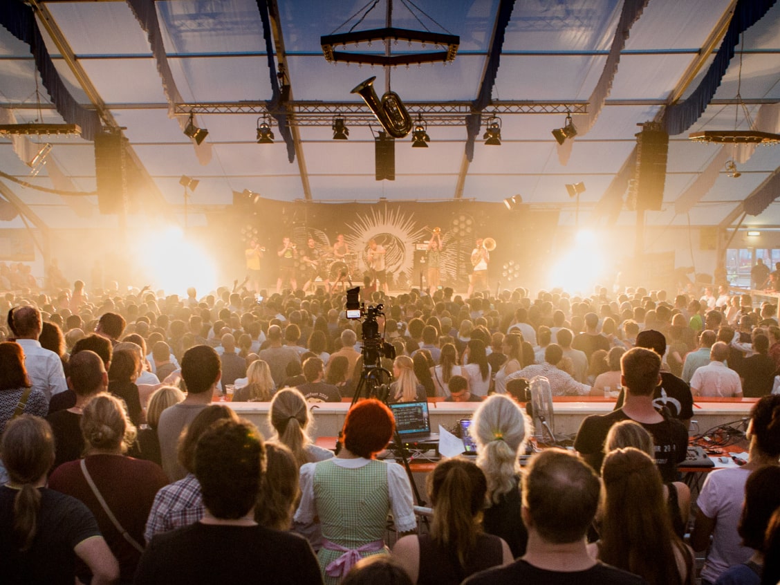 A large crowd watches a band perform on stage in a tent, with bright lights and sound equipment visible.