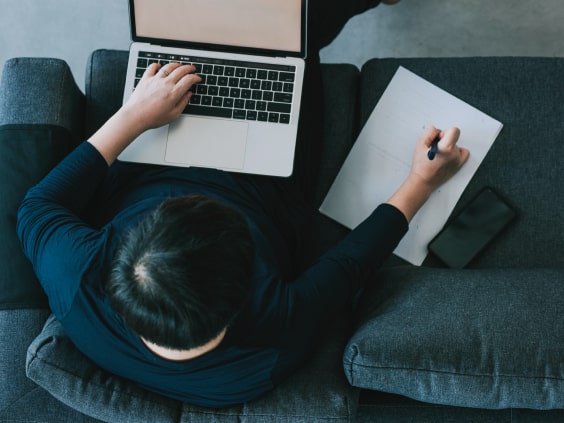 Person on a couch using a laptop and writing on paper, with a phone beside them.