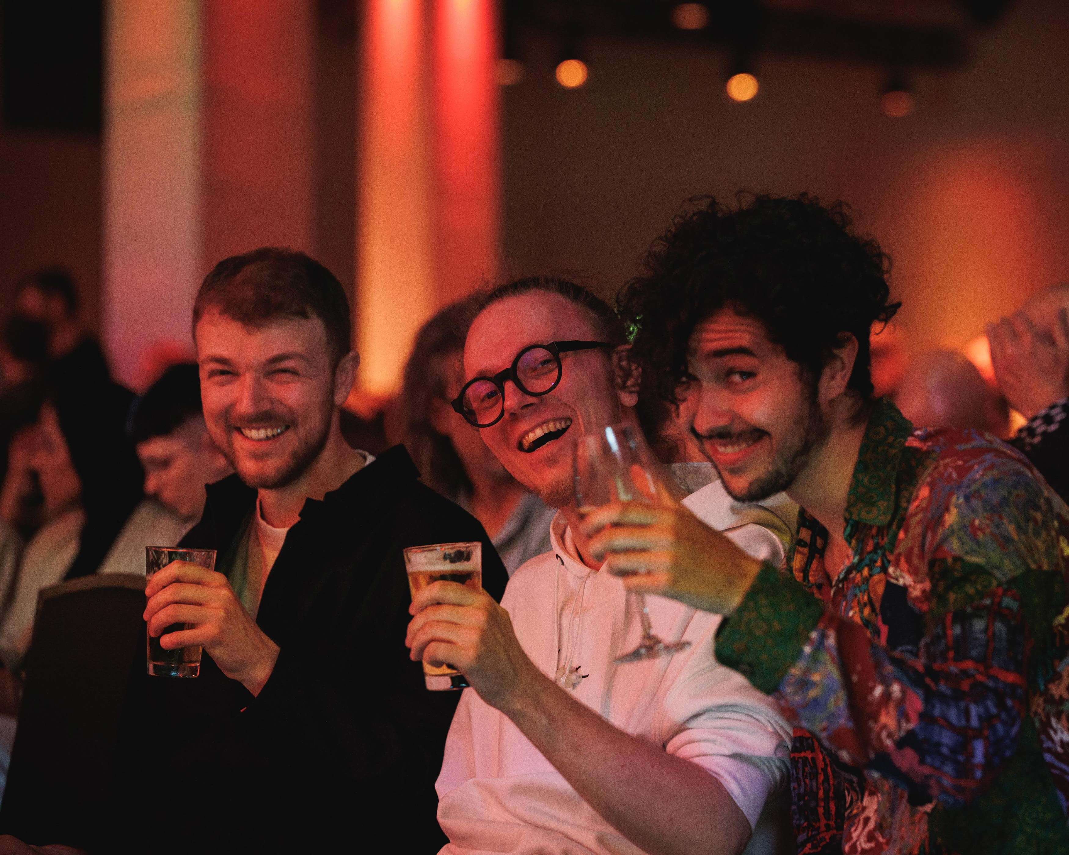 Three smiling people holding drinks, seated in a dimly lit room with colorful lights in the background.