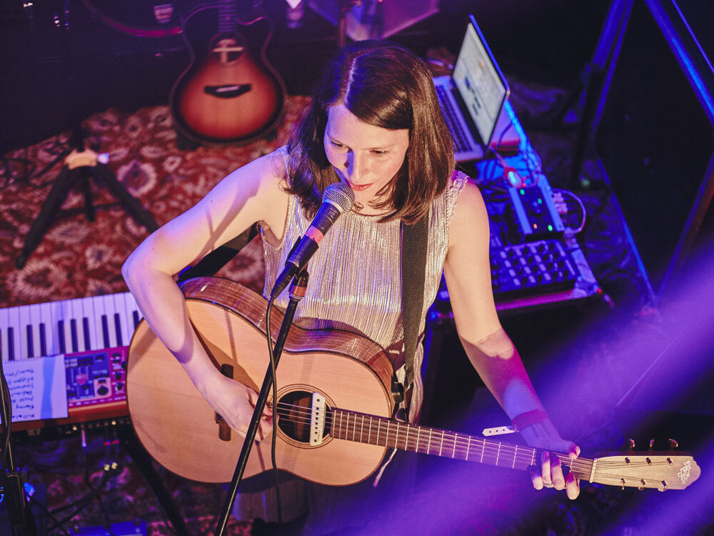 Musikerin spielt Gitarre auf Bühne, umgeben von Keyboard und Laptop, in lila Licht getaucht.
