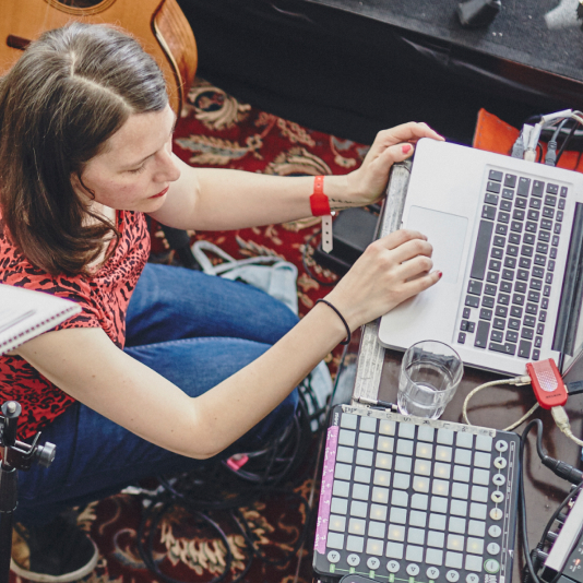 A person in a red shirt uses a laptop on a patterned rug, surrounded by musical equipment and a glass of water.