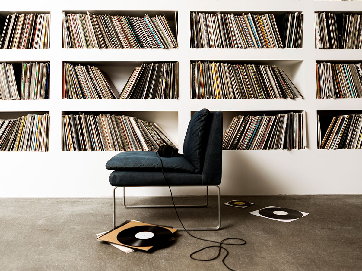 A wall of vinyl records, a blue chair, and headphones with records scattered on the floor.