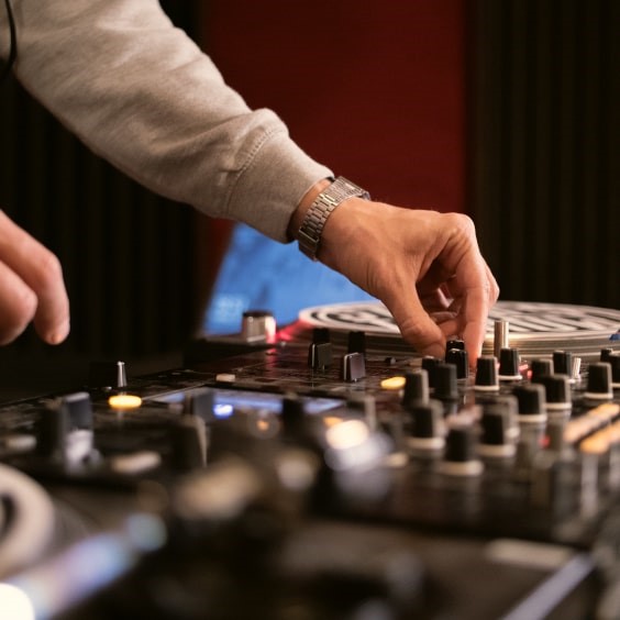 A person adjusts knobs on a DJ mixer, wearing a gray sweatshirt and watch, with a blurred background.
