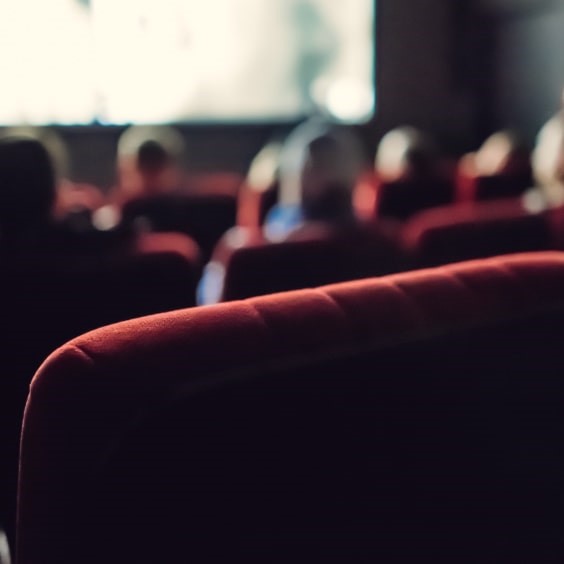 Blurred image of people sitting in red theater seats facing a large screen showing a movie.