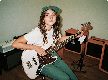 Eine junge Frau mit grüner Kappe spielt im sitzen Gitarre und blickt in die Kamera.