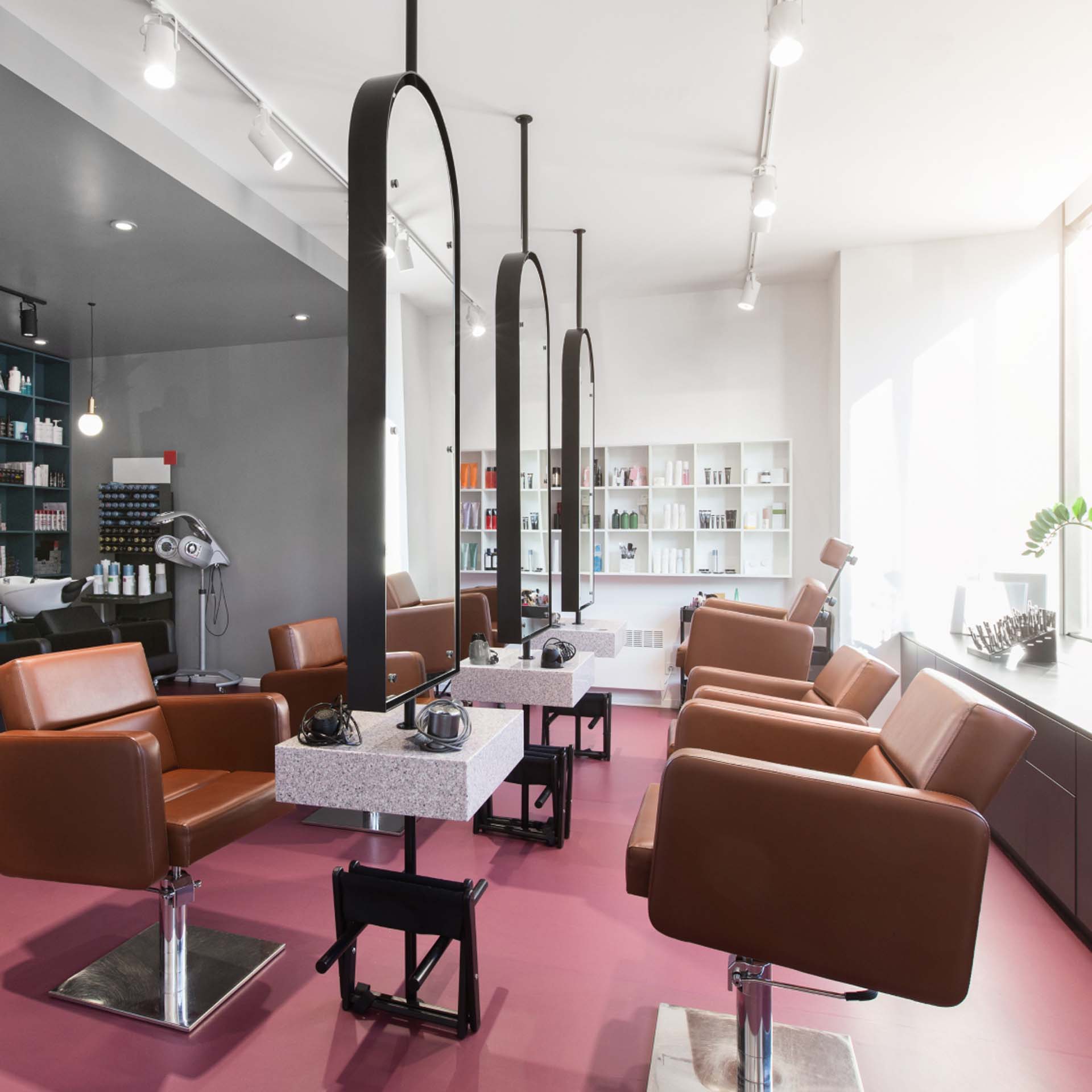 Modern salon with brown chairs, pink floor, large mirrors, and shelves of hair products under bright lighting.