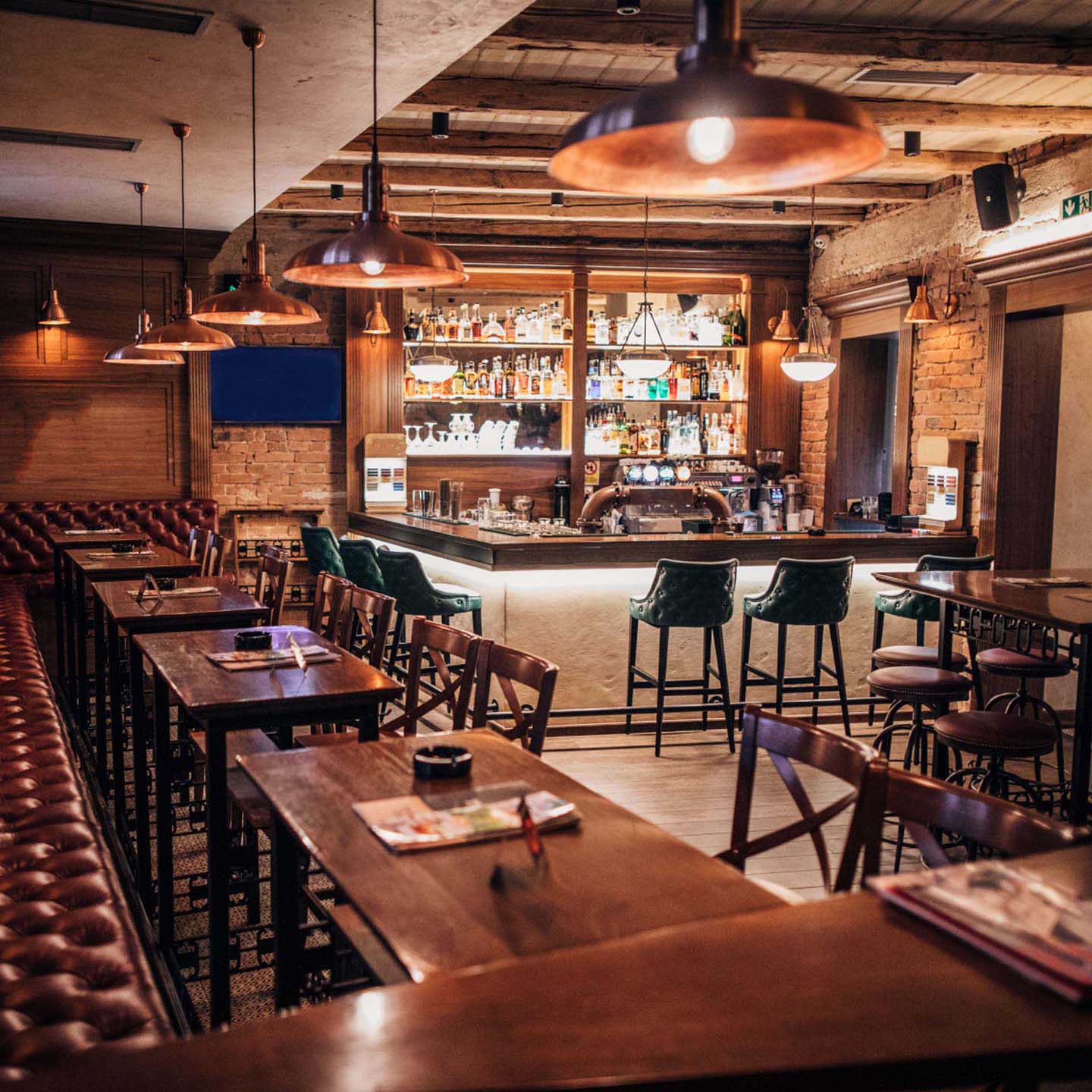 Cozy bar with wooden tables, leather chairs, and a well-stocked backlit bar counter under warm pendant lights.