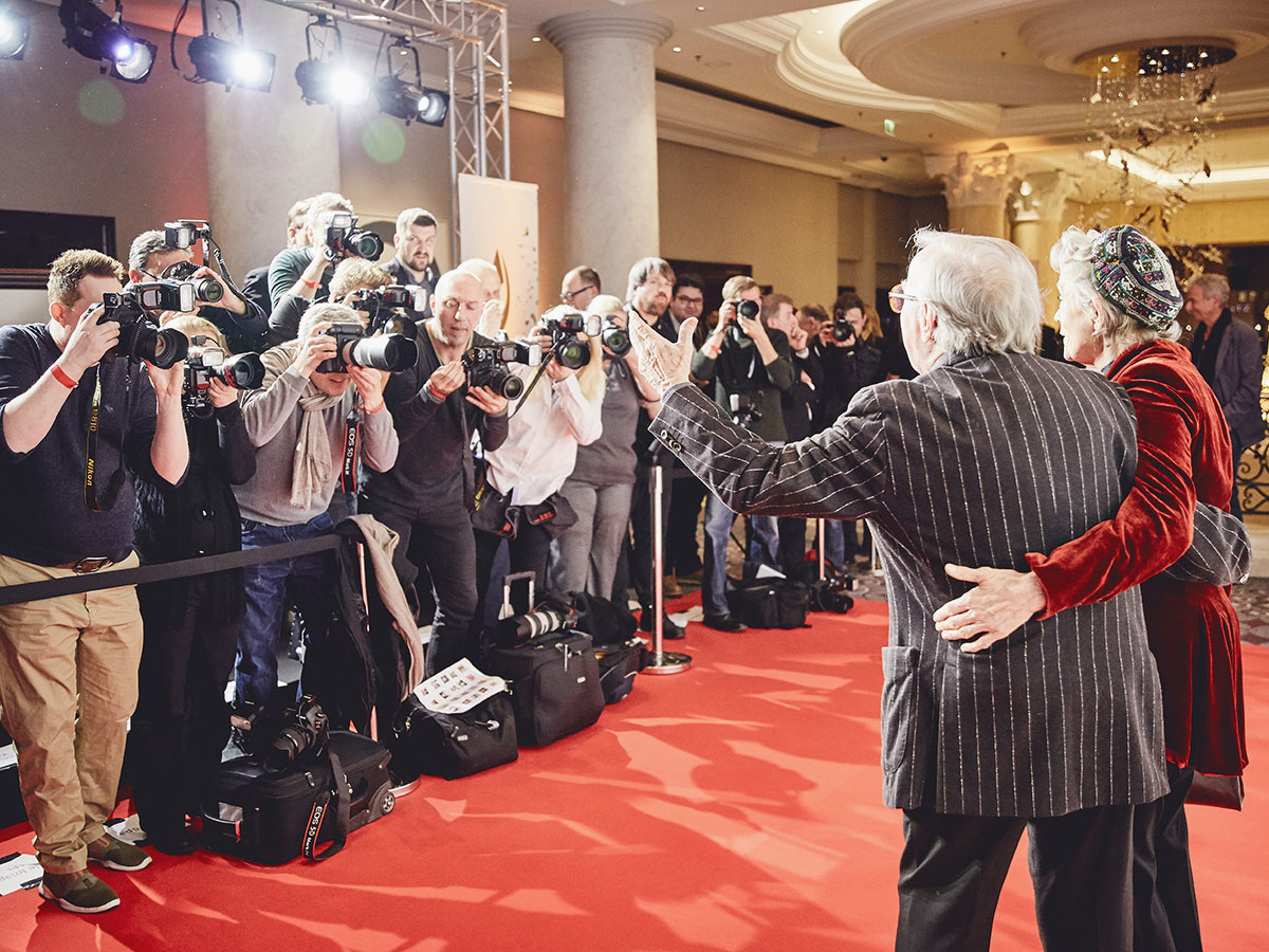 Two people pose on a red carpet, facing photographers with cameras and bright lights in a crowded indoor setting.
