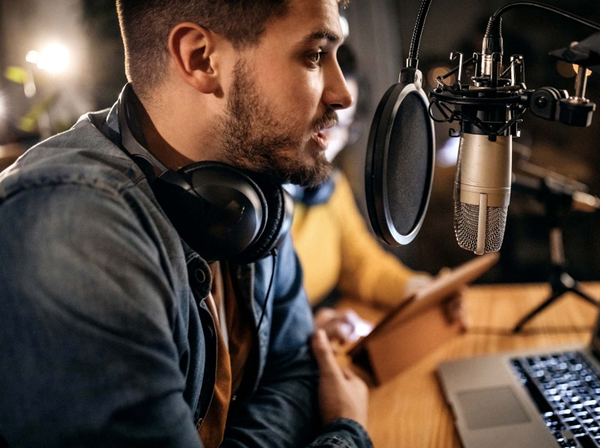 Man with headphones speaks into a microphone at a desk with a laptop, another person in the background holds a tablet.