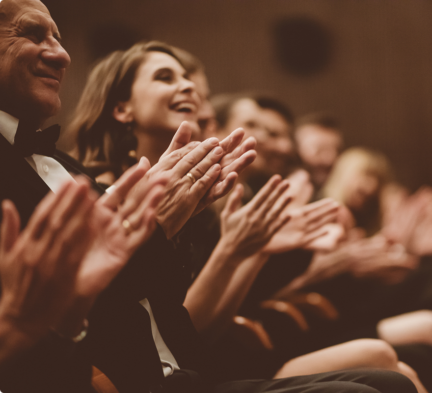Audience clapping in a theater, with people in formal attire smiling and applauding enthusiastically.