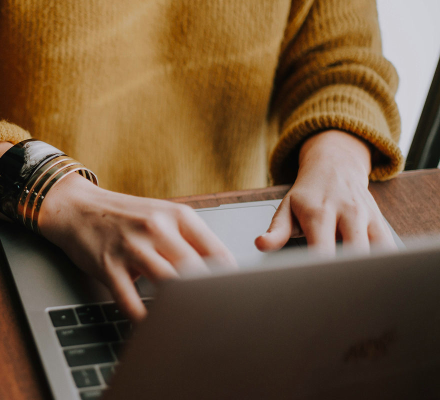 Person in a yellow sweater typing on a laptop, wearing multiple bracelets.