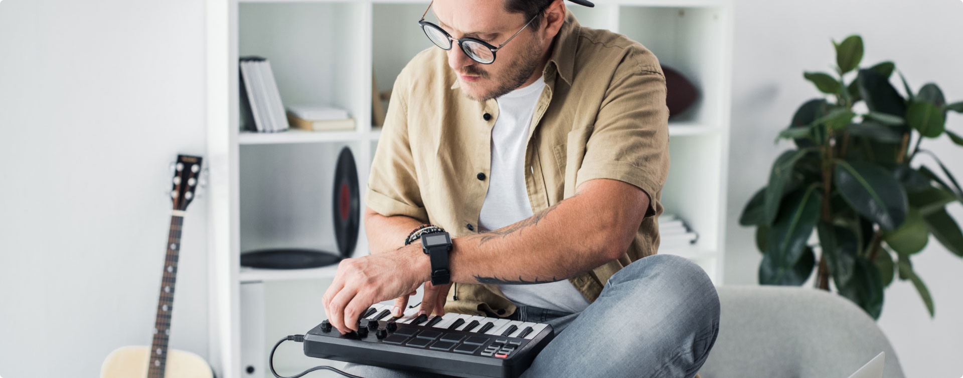 Man in glasses using a MIDI controller on sofa, with a guitar and plant in the background.
