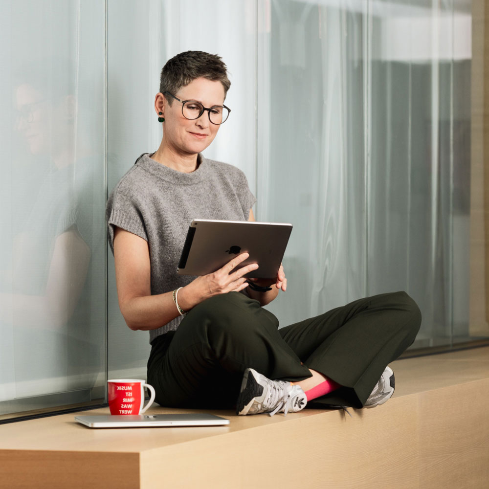 A person with short hair and glasses sits on a ledge, using a tablet. A red mug and laptop are beside them.