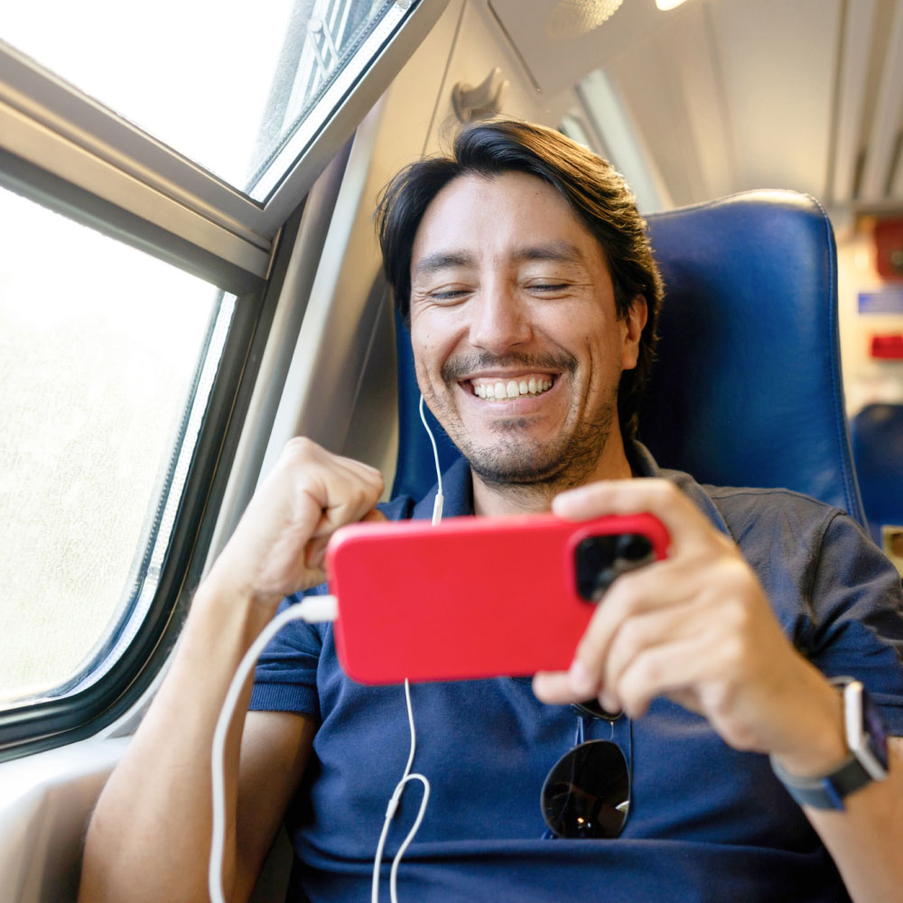 Man smiling on a train, wearing earphones, holding a red phone, and sitting by the window.