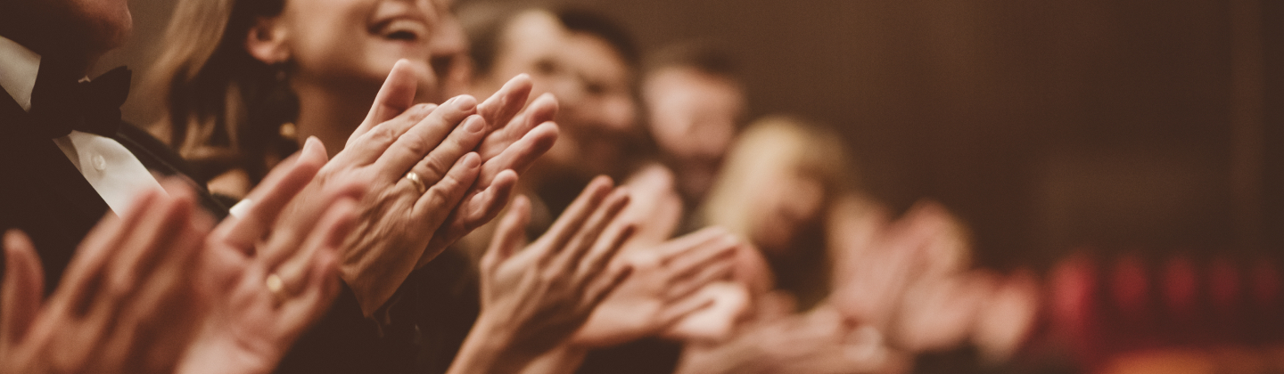 Audience clapping in a theater, with people in formal attire smiling and applauding enthusiastically.