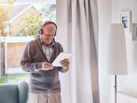 Ein älterer Mann mit Kopfhörern schaut auf ein Tablet, stehend neben einem Vorhang und einer Stehlampe.