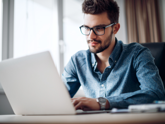 A person in glasses and a blue shirt works on a laptop at a desk in a bright room.