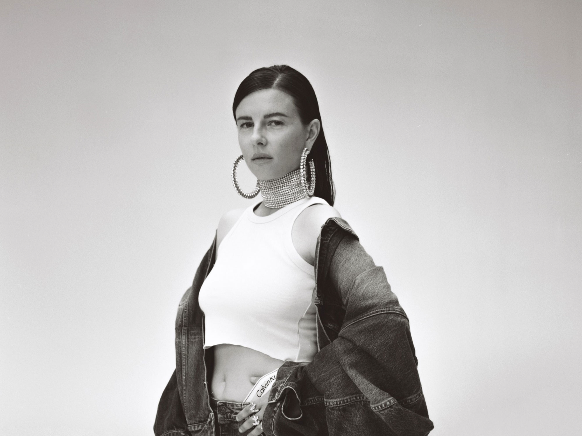 Woman in a white crop top with large hoop earrings and denim jacket poses against a plain background.