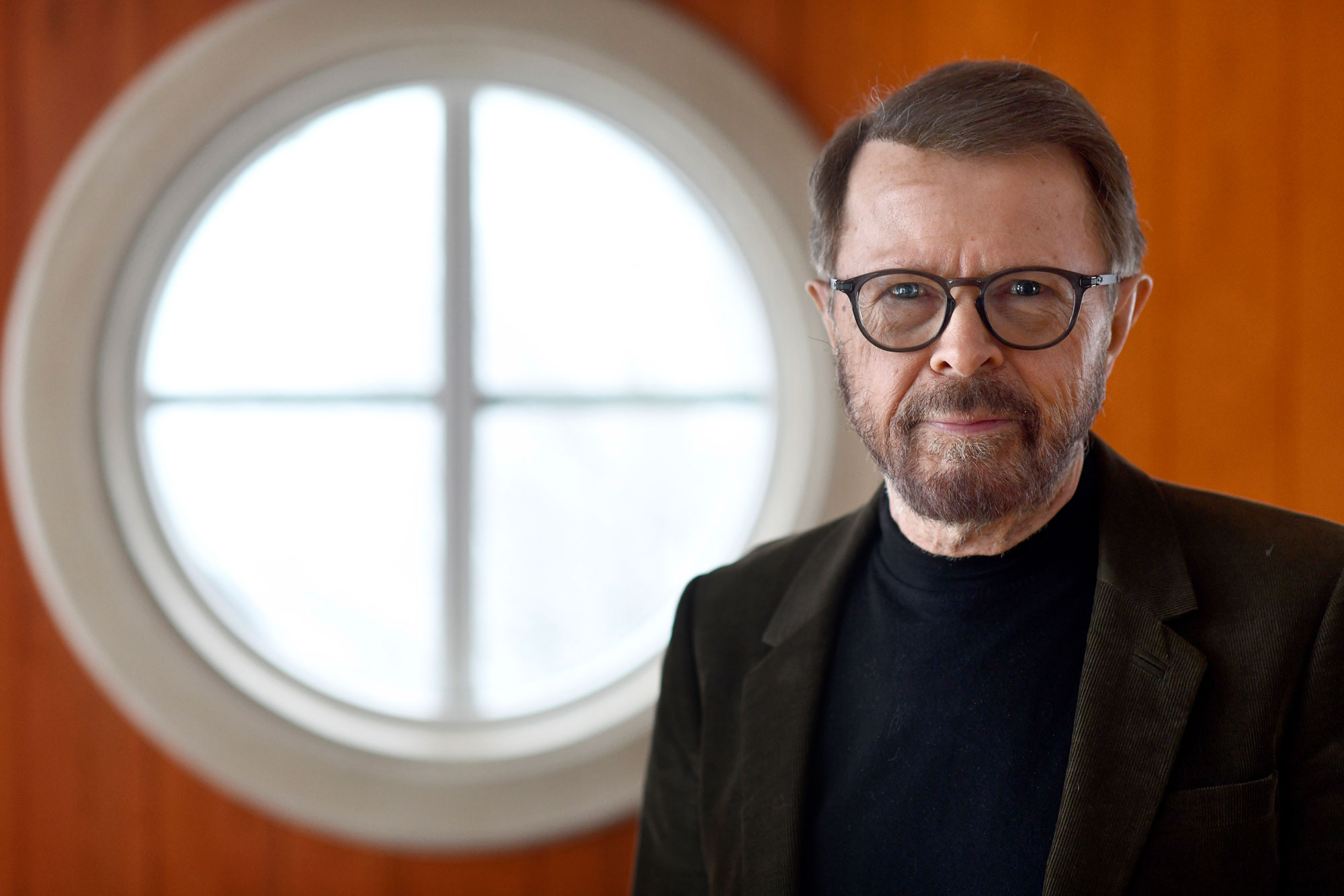 A person with glasses stands in front of a round window, wearing a black shirt and dark blazer, with an orange wall background.