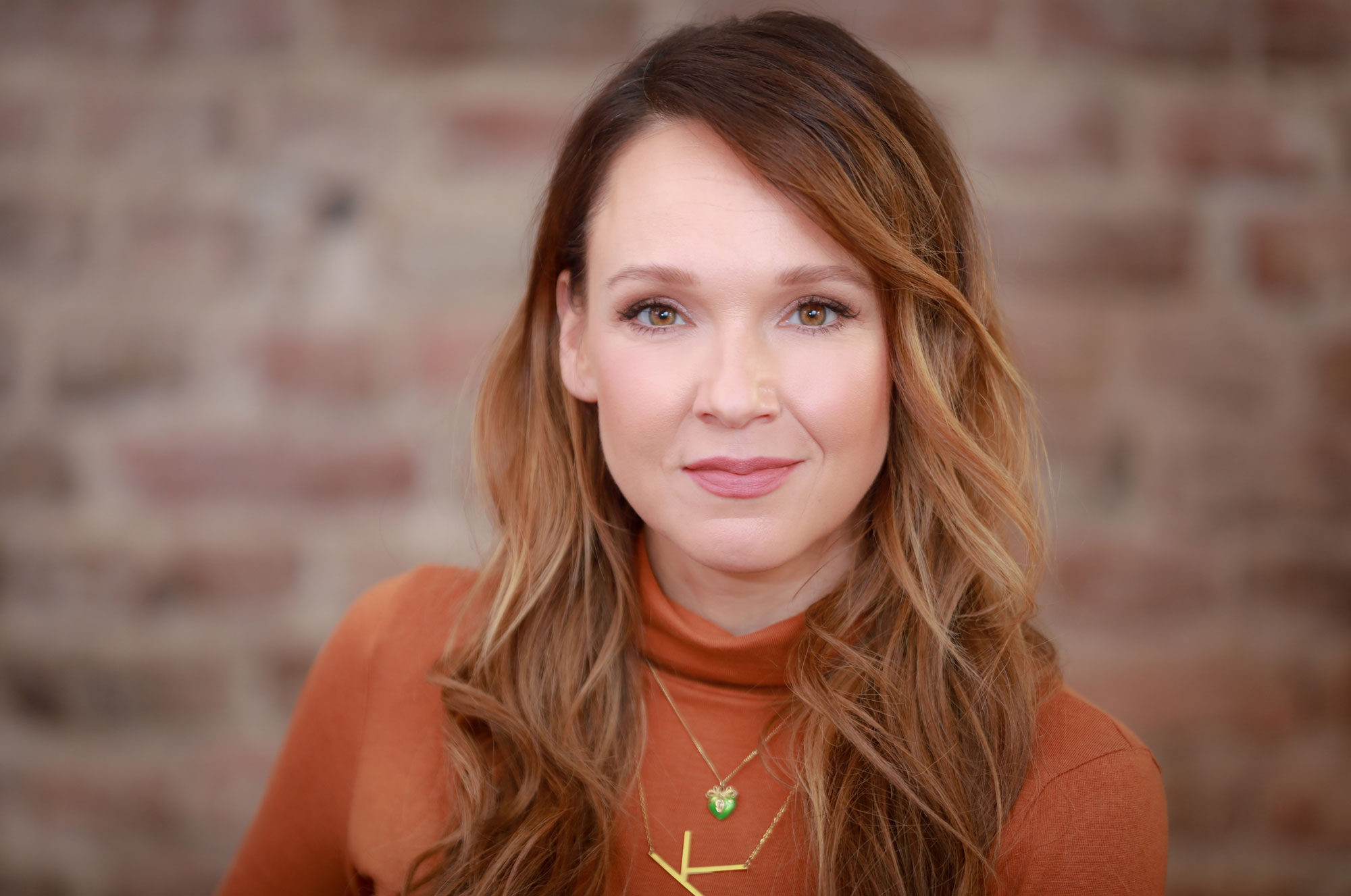 Woman with long brown hair in orange top, smiling against a blurred brick wall background.