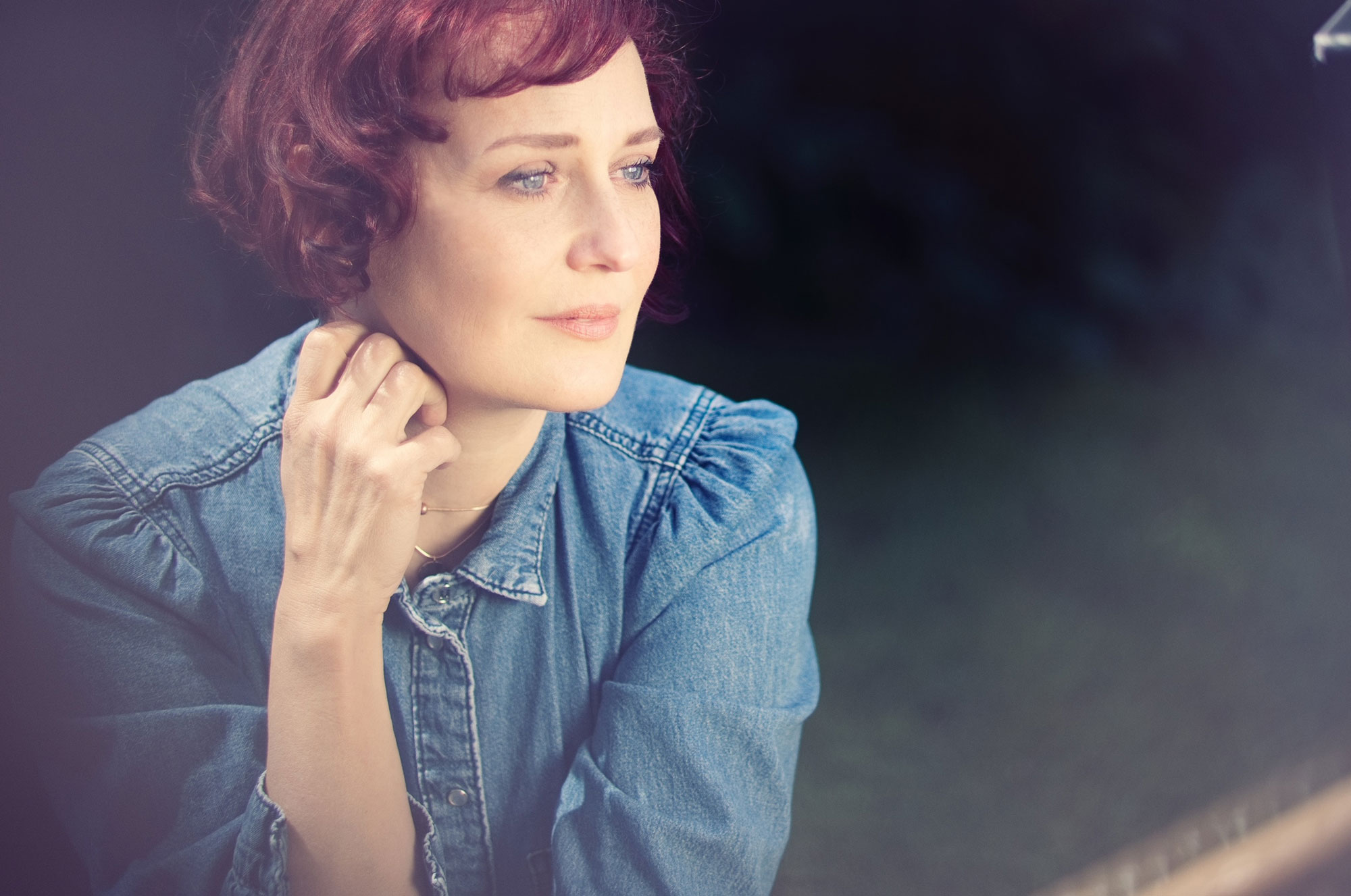 Woman with red hair in a denim shirt gazes thoughtfully out a window, hand resting on her chin.