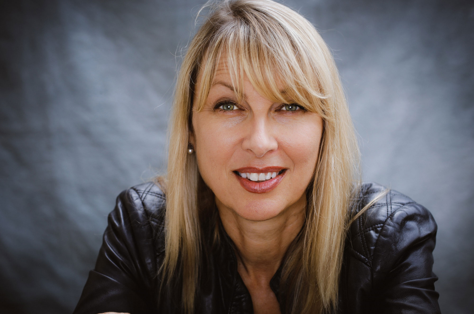 Smiling woman with blonde hair in a black leather jacket against a gray background.