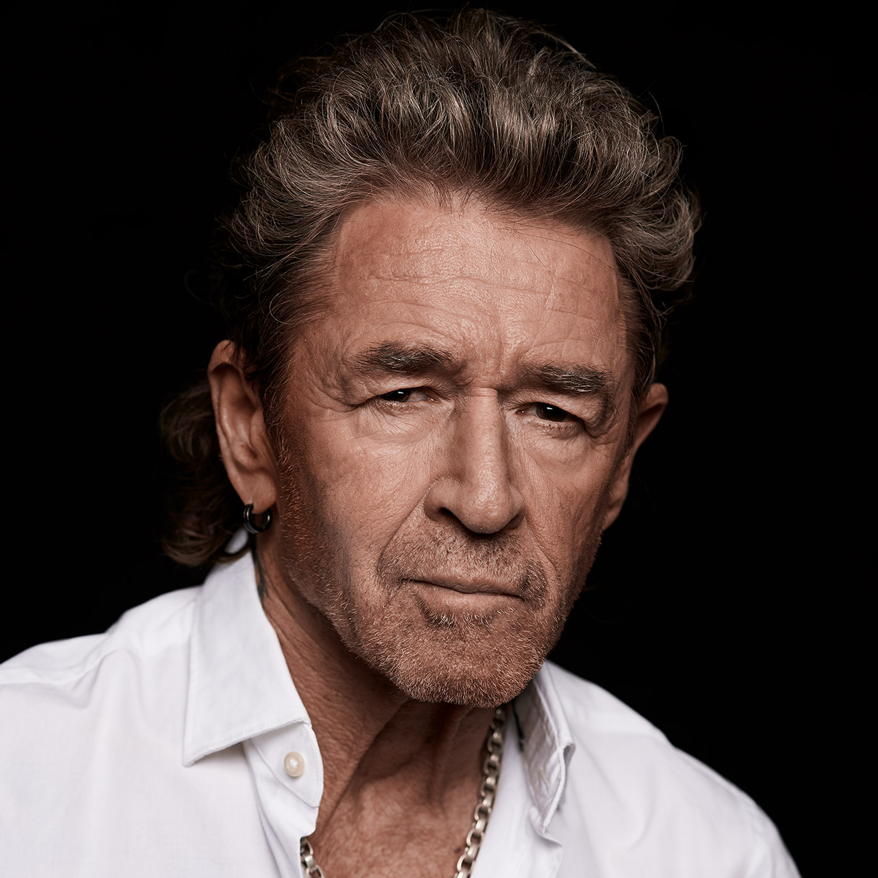 Elderly man with gray hair, wearing a white shirt and necklace, stares intently against a dark background.