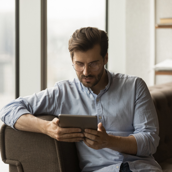 Man in glasses sitting on a couch, reading a tablet near a large window.