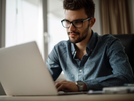 A man with glasses works on a laptop at a desk in a bright room.