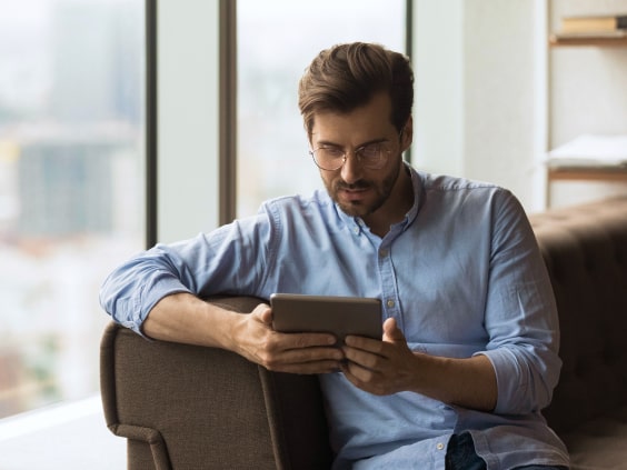 Man in a blue shirt sits on a couch, reading a tablet by a large window.