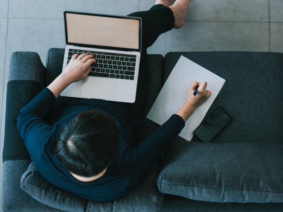 Person on a gray sofa using a laptop with a notepad and phone beside them.