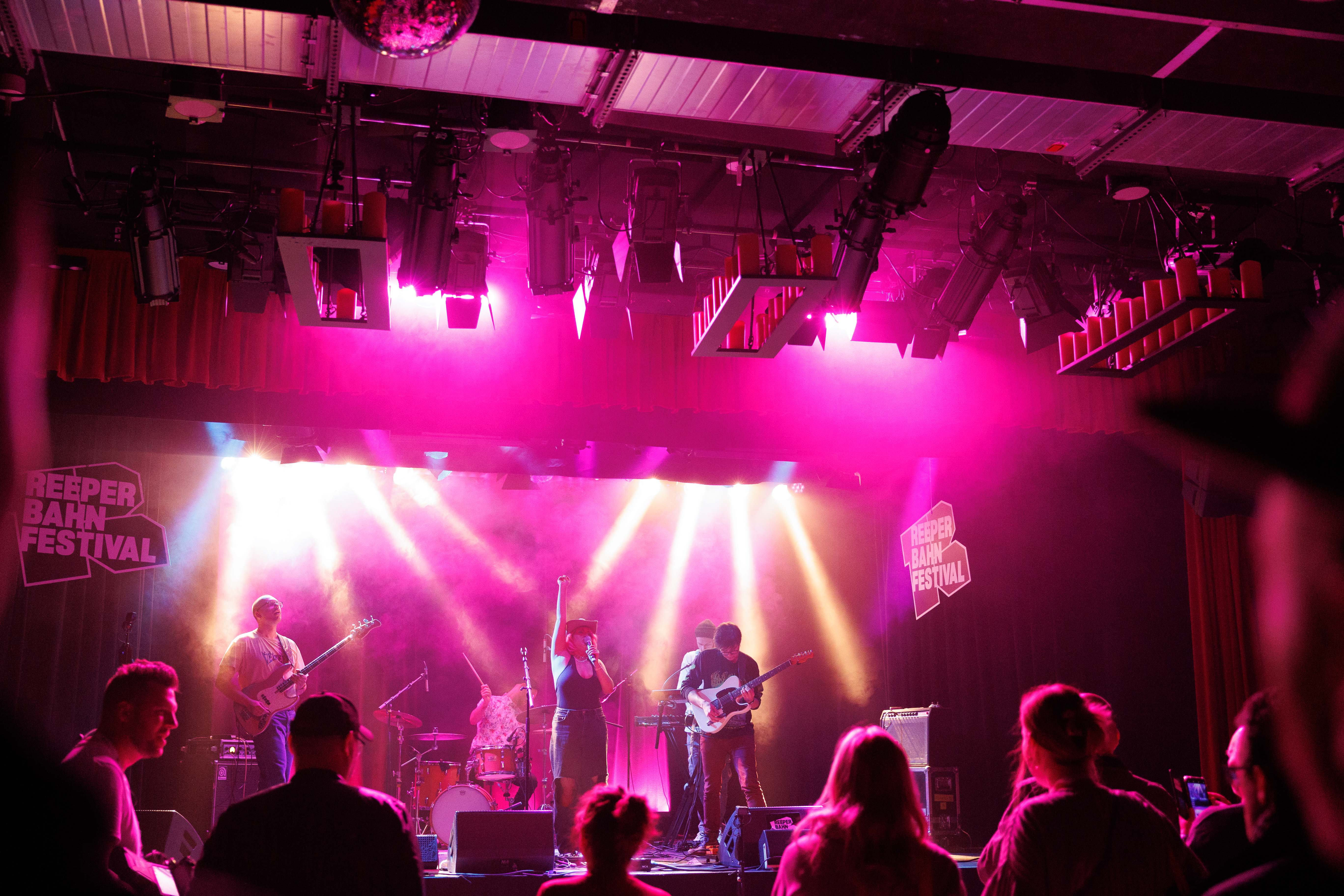 Eine Band spielt auf einer Bühne mit pinkem Licht beim Reeperbahn Festival, Zuschauer im Vordergrund.