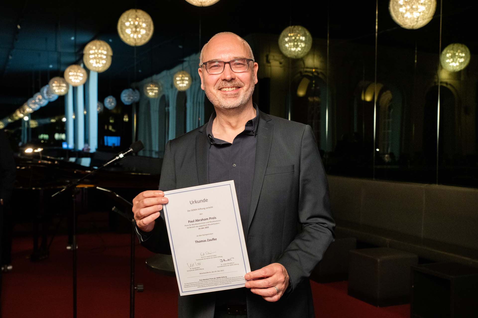 Ein lächelnder Mann hält eine Urkunde in einem eleganten Raum mit glänzenden Kugelleuchten.