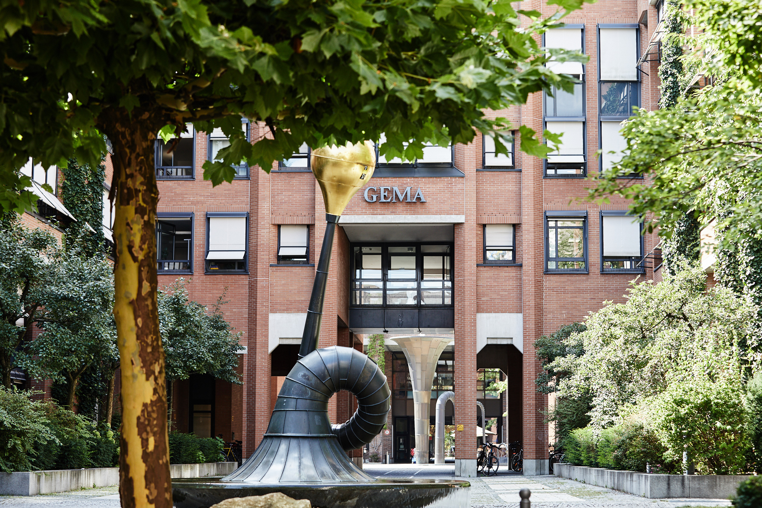 Skulptur mit goldener Spitze vor Backsteingebäude, umgeben von Bäumen und Pflanzen.