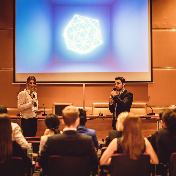 Two presenters with microphones stand in front of an audience, under a screen displaying a geometric shape.