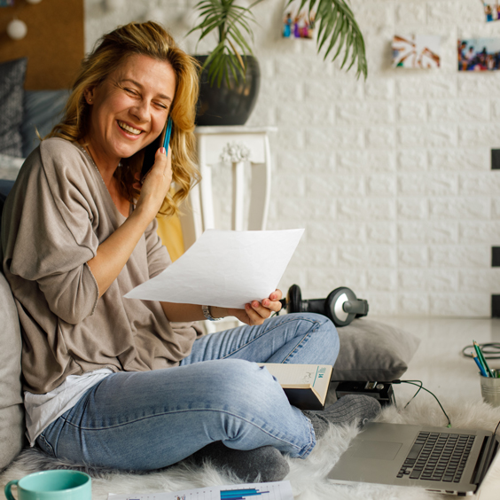 A woman sitting on a couch smiles while talking on the phone, holding a paper, with a laptop and headphones nearby.