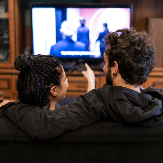 A couple sits on a couch watching TV, with one person pointing at the screen, showing blurred figures in suits.