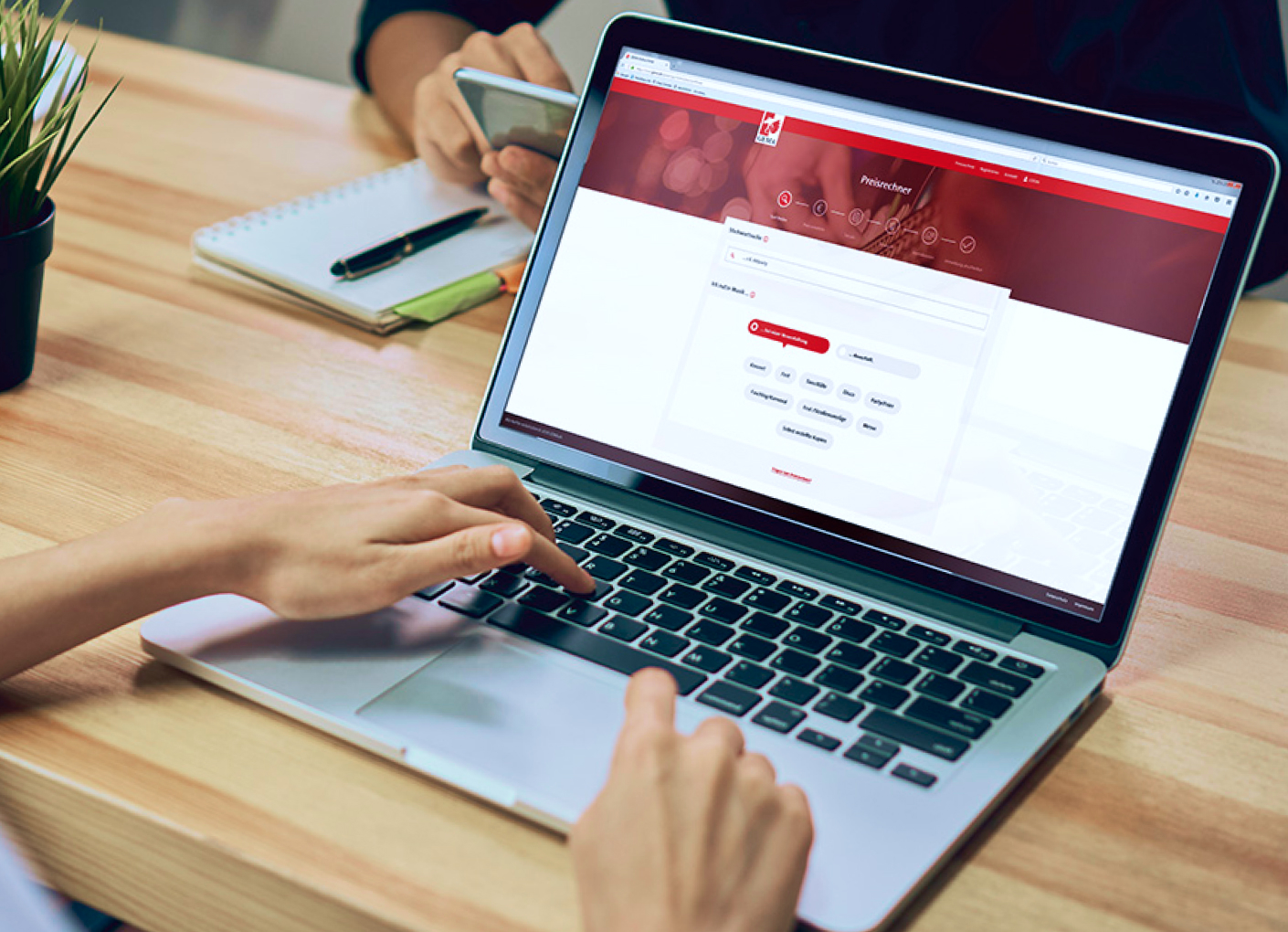 Hands typing on a laptop with a red website open, next to a notebook, pen, and smartphone on a wooden desk.