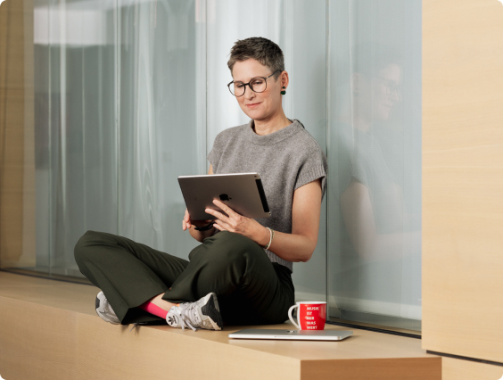 A person sits cross-legged on a ledge, using a tablet. A red mug and closed laptop are beside them.