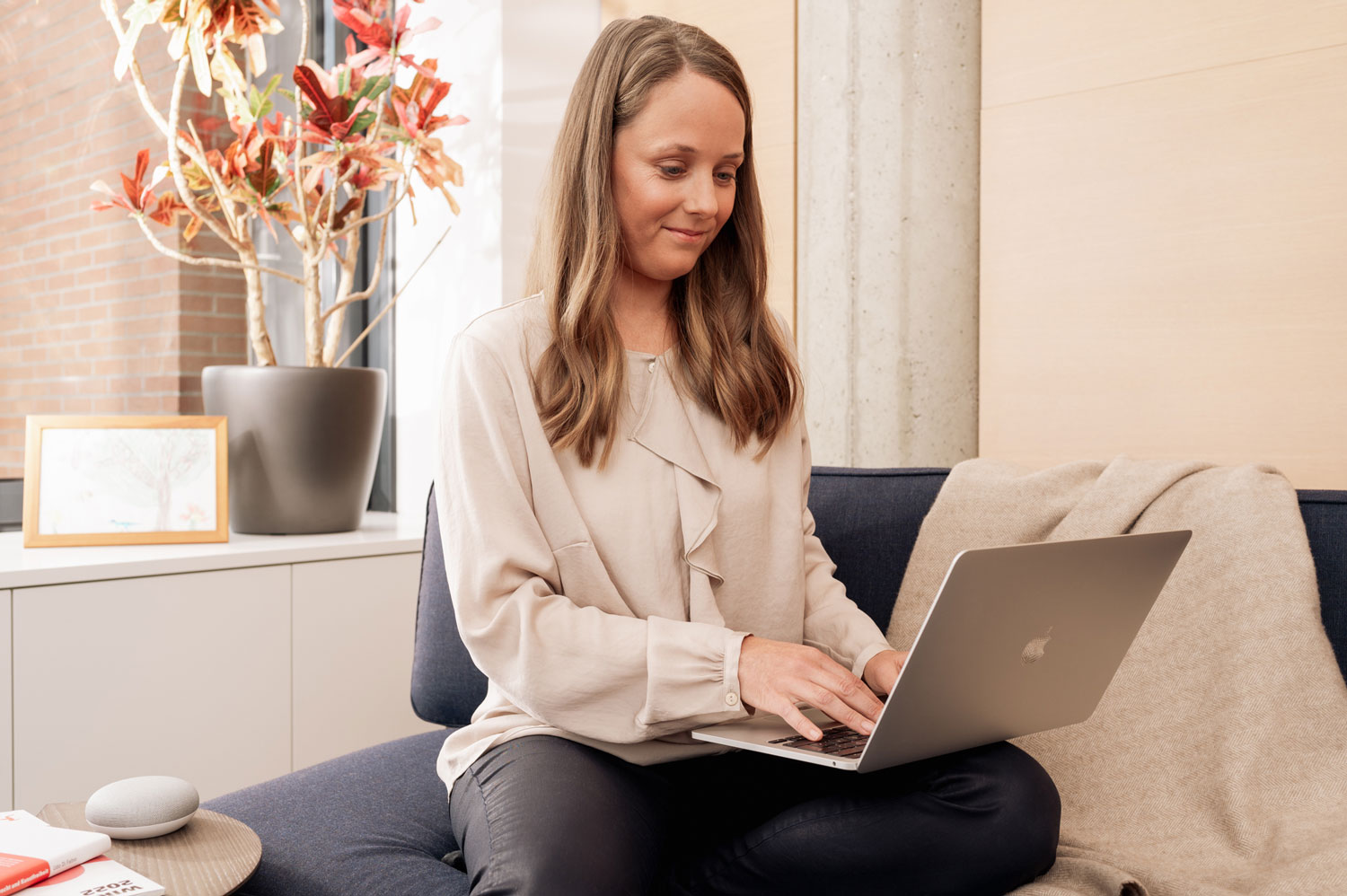 Woman in beige blouse using a laptop on a sofa, with a potted plant and framed picture in the background.
