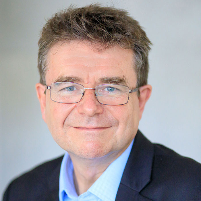 A man with glasses and a suit smiles against a plain background.