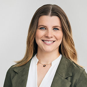 A smiling woman with long hair wearing a white blouse and green jacket against a plain background.